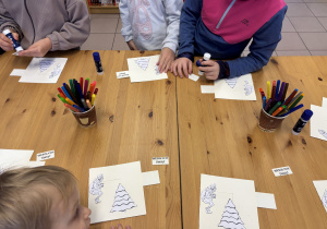 Dzieci stoją przy stole i ozdabiają świąteczne kartki.