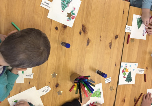 Dzieci stoją przy stole i ozdabiają świąteczne kartki.