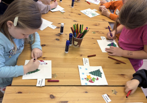 Dzieci stoją przy stole i ozdabiają świąteczne kartki.