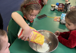 Dzieci siedzą przy stole, dziewczynka wsypuje mąkę do miski.
