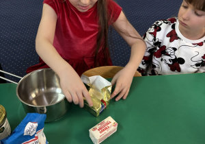 Dzieci siedzą przy stole, dziewczynka rozwija opakowanie z masłem.