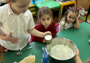 Dzieci siedzą przy stole, chłopiec wsypuje cukier do miski.