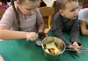 Dzieci siedzą przy stole, dziewczynka wkłada miód do rondelka.