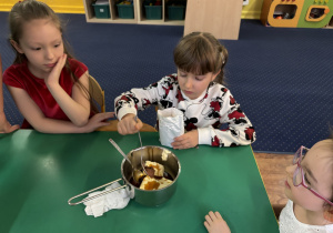 Dzieci siedzą przy stole, dziewczynka wsypuje kakao do rondelka.