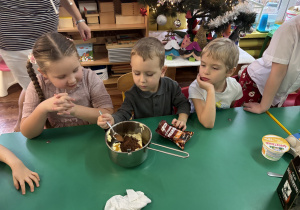 Dzieci siedzą przy stole, chłopiec wsypuje przyprawę do piernika do rondelka.