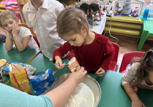 Dzieci siedzą przy stole, dziewczynka wsypuje proszek do pieczenia do miski.
