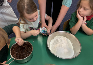 Dzieci siedzą przy stole, dziewczynka wsypuje sodę do miski.