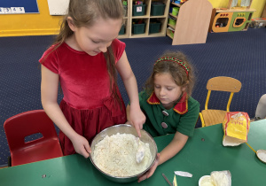 Dzieci siedzą przy stole, dziewczynka miesza produkty w misce.