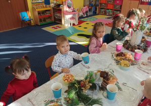 dzieci świętują wigilię klasową, siedzą przy stolikach, na których widać słodycze, owoce, soki