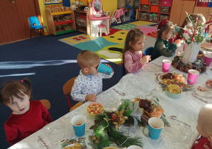 dzieci świętują wigilię klasową, siedzą przy stolikach, na których widać słodycze, owoce, soki