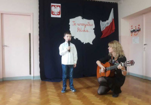 Chłopiec recytuje wiersz, nauczyciel przygrywa mu na gitarze.