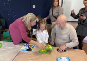 Dzieci wraz z dziadkami siedzą przy stołach i wykonują prezenty.
