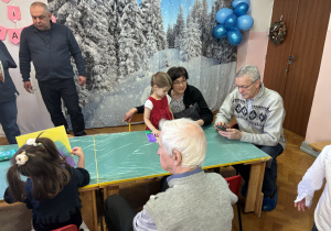 Dzieci wraz z dziadkami siedzą przy stołach i wykonują prezenty.