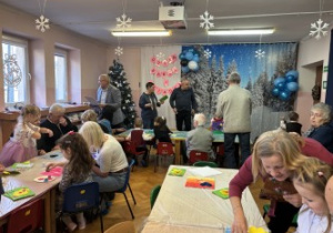 Dzieci wraz z dziadkami siedzą przy stołach i wykonują prezenty.