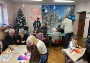 Dzieci wraz z dziadkami siedzą przy stołach i wykonują prezenty.