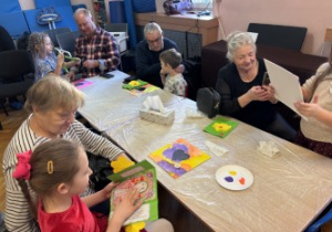 Dzieci wraz z dziadkami siedzą przy stołach i wykonują prezenty.