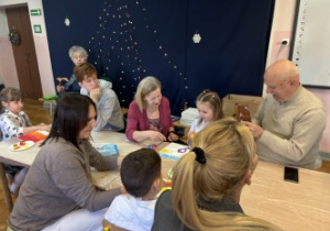 Dzieci wraz z dziadkami siedzą przy stołach i wykonują prezenty.