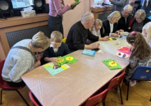 Dzieci wraz z dziadkami siedzą przy stołach i wykonują prezenty.