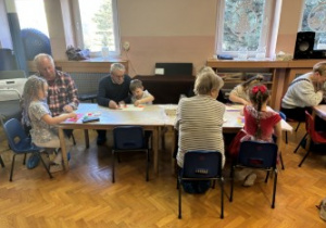 Dzieci wraz z dziadkami siedzą przy stołach i wykonują prezenty.