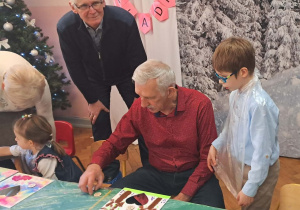 Dzieci wraz z dziadkami siedzą przy stołach i wykonują prezenty.