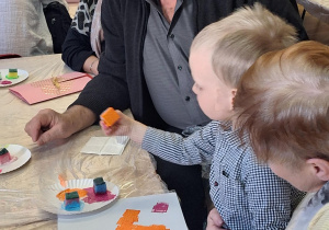 Dzieci wraz z dziadkami siedzą przy stołach i wykonują prezenty.