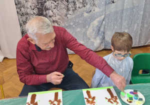 Dzieci wraz z dziadkami siedzą przy stołach i wykonują prezenty.