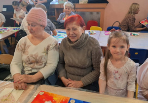 Dzieci wraz z dziadkami siedzą przy stołach i wykonują prezenty.