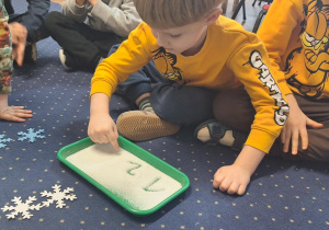 Chłopiec siedzi na dywanie i pisze cyfry na piasku.