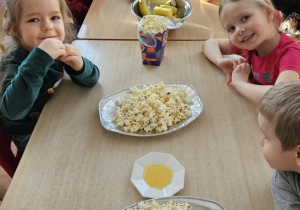 Dzieci siedzą przy stole na którym stoi popcorn.