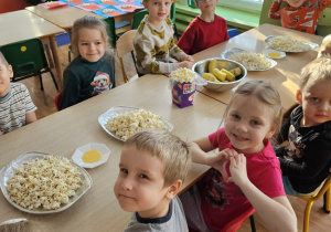 Dzieci siedzą przy stole na którym stoi popcorn.
