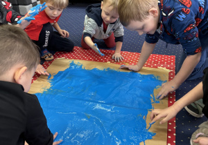 Dzieci na dywanie malują karton niebieską farbą używając dłoni.