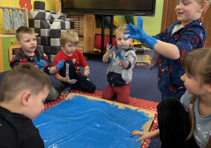 Dzieci na dywanie malują karton niebieską farbą używając dłoni.