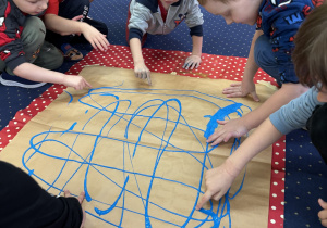 Dzieci na dywanie malują karton niebieską farbą używając dłoni.