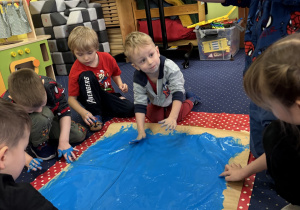 Dzieci na dywanie malują karton niebieską farbą używając dłoni.