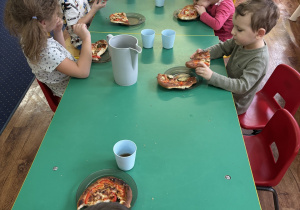 Dzieci siedzą przy stolikach i zjadają pizzę.