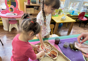 Dzieci siedzą przy stolikach i przygotowują własną pizzę.