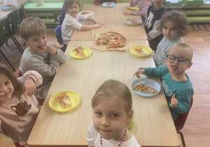 Dzieci siedzą przy stole na którym jest upieczona pizza.