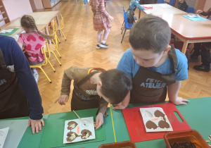 dzieci ubrane w brązowe fartuszki biorą udział w czekoladowych warsztatach, samodzielnie ozdabiają czekoladę w kształcie serca