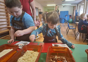dzieci ubrane w brązowe fartuszki biorą udział w czekoladowych warsztatach, samodzielnie ozdabiają czekoladę w kształcie serca