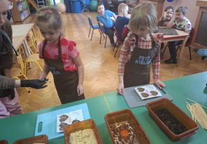 dzieci ubrane w brązowe fartuszki biorą udział w czekoladowych warsztatach, samodzielnie ozdabiają czekoladę w kształcie serca