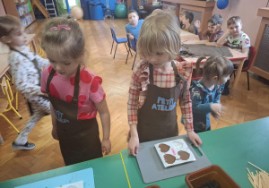 dzieci ubrane w brązowe fartuszki biorą udział w czekoladowych warsztatach, samodzielnie ozdabiają czekoladę w kształcie serca