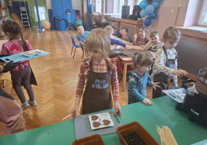 dzieci ubrane w brązowe fartuszki biorą udział w czekoladowych warsztatach, samodzielnie ozdabiają czekoladę w kształcie serca