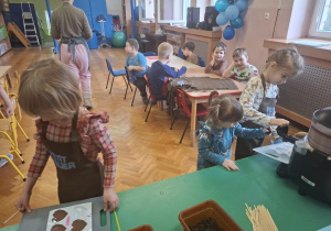 dzieci ubrane w brązowe fartuszki biorą udział w czekoladowych warsztatach, samodzielnie ozdabiają czekoladę w kształcie serca