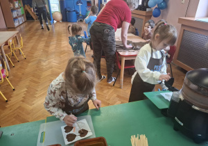 dzieci ubrane w brązowe fartuszki biorą udział w czekoladowych warsztatach, samodzielnie ozdabiają czekoladę w kształcie serca