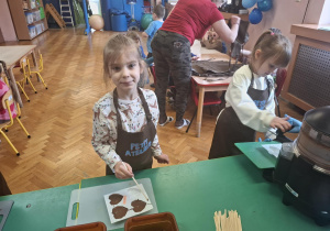 dzieci ubrane w brązowe fartuszki biorą udział w czekoladowych warsztatach, samodzielnie ozdabiają czekoladę w kształcie serca