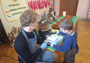 dzieci ubrane w brązowe fartuszki biorą udział w czekoladowych warsztatach, samodzielnie ozdabiają czekoladę w kształcie serca