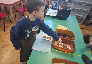 dzieci ubrane w brązowe fartuszki biorą udział w czekoladowych warsztatach, samodzielnie ozdabiają czekoladę w kształcie serca