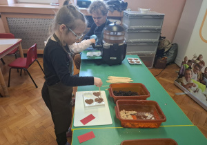 dzieci ubrane w brązowe fartuszki biorą udział w czekoladowych warsztatach, samodzielnie ozdabiają czekoladę w kształcie serca