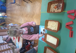 dzieci ubrane w brązowe fartuszki biorą udział w czekoladowych warsztatach, samodzielnie ozdabiają czekoladę w kształcie serca