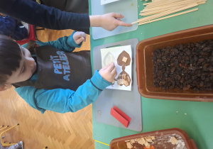 dzieci ubrane w brązowe fartuszki biorą udział w czekoladowych warsztatach, samodzielnie ozdabiają czekoladę w kształcie serca